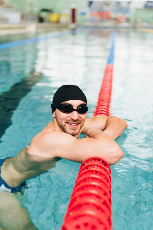 A Smiling Male Swimmer In A Pool, Resting On A Lane Line. The Individual Is Wearing A Black Swim Cap And Goggles.