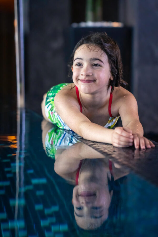 A Young Girl With Wet Hair, Wearing A Green And White Swimsuit With Red Straps, Is Smiling While Lying On The Edge Of A Swimming Pool