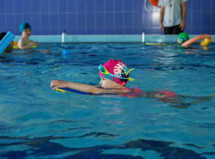 A Young Girl In A Pink Cap And Goggles Uses A Kickboard During A Swimming Lesson