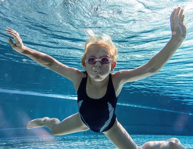 A Young Girl With Blonde Hair And Pink Swimming Goggles Is Swimming Underwater In A Pool, Likely During A Swimming Lesson Or Activity.
